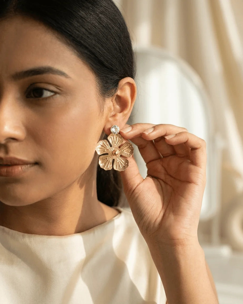 Model wearing golden coloured metal flower shape earrings with tetured on it , a diamond on top part of earrings