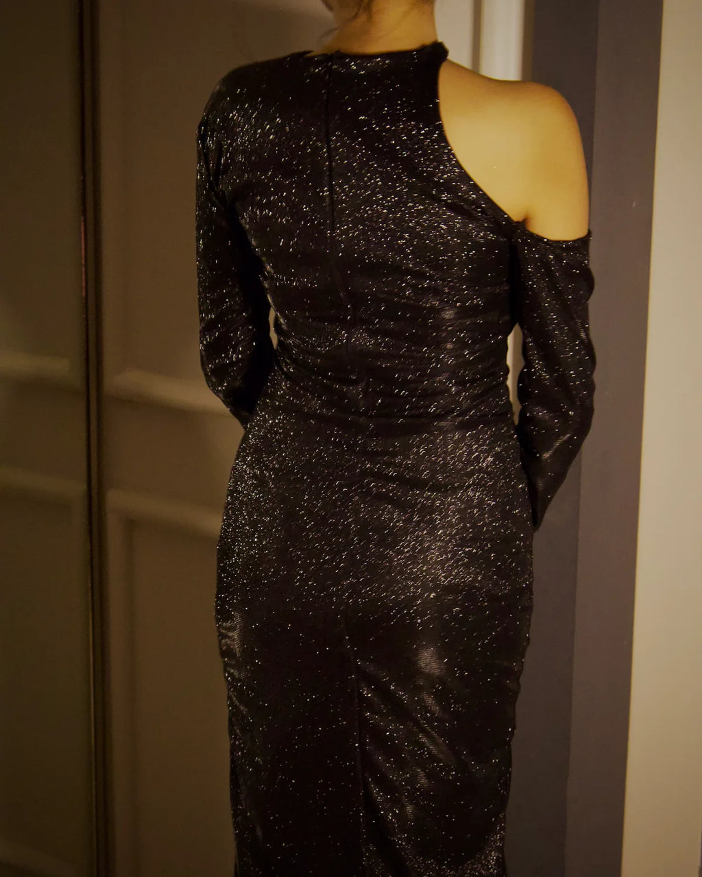 A model wearing a sparkling black one-shoulder evening gown with a cold-shoulder cut, shown from the back in an evening a beautiful glitter dress.