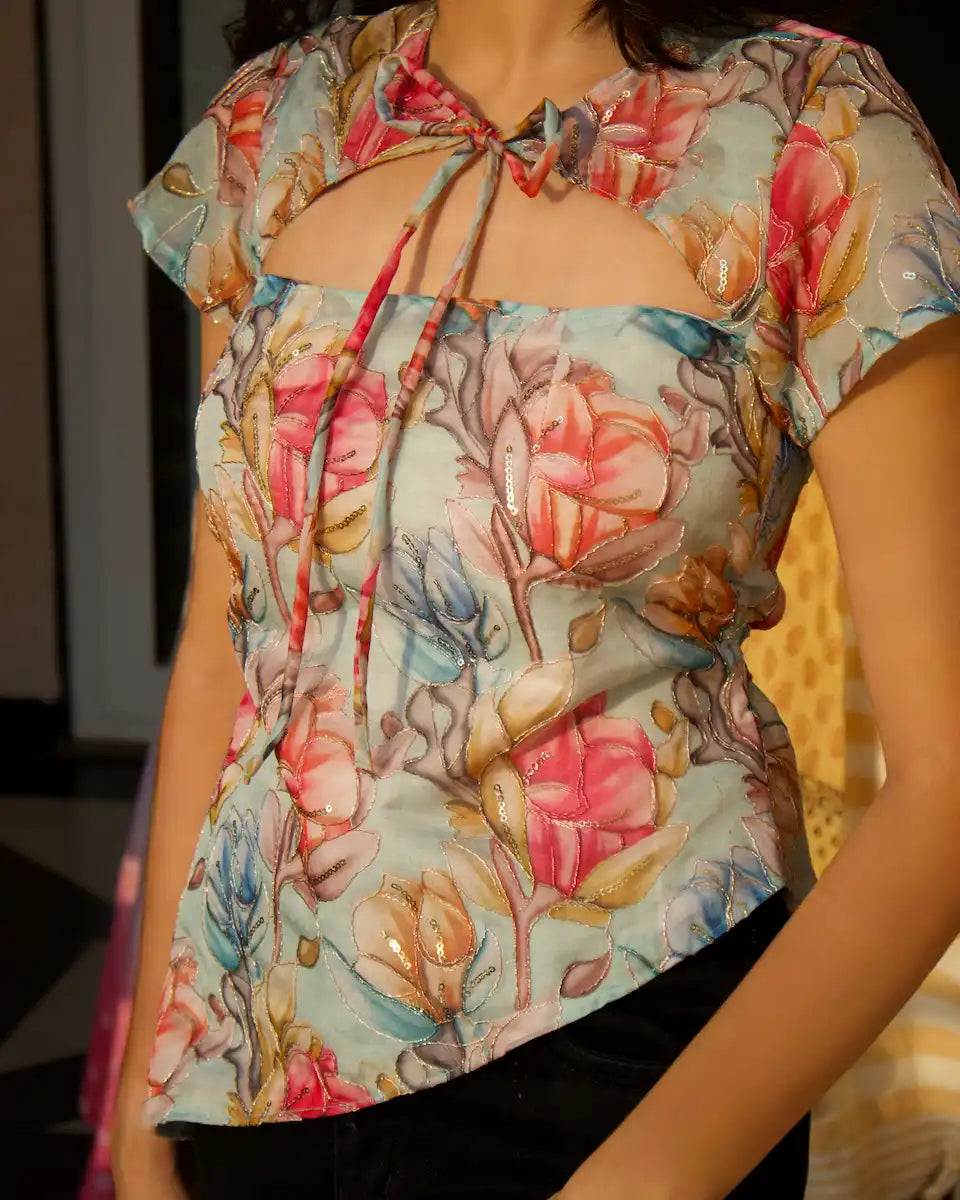 A woman wearing a short-sleeve blouse with a floral print, a square neckline, and a front tie detail, paired with black bottoms.