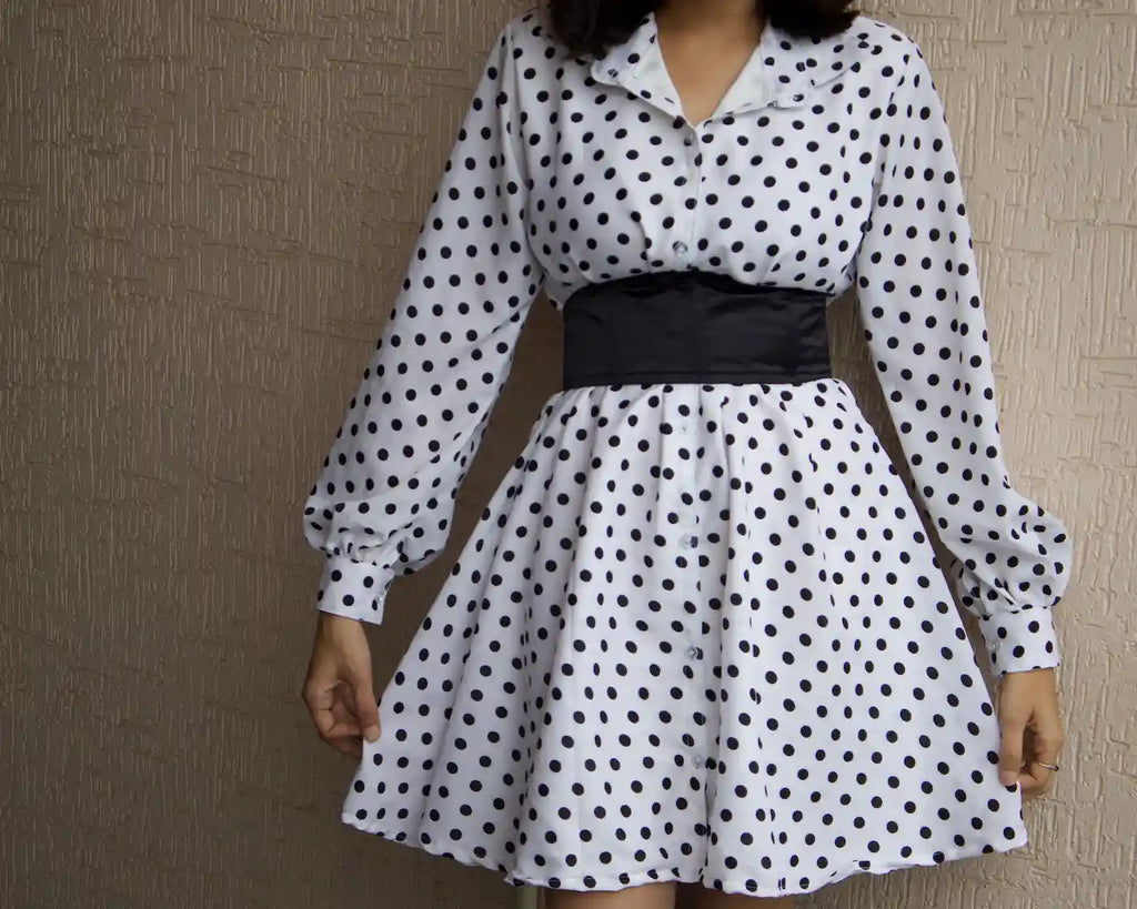 A woman wearing a white long-bell sleeve dress with black polka dots, featuring a wide black corset  waistband, with aline flare